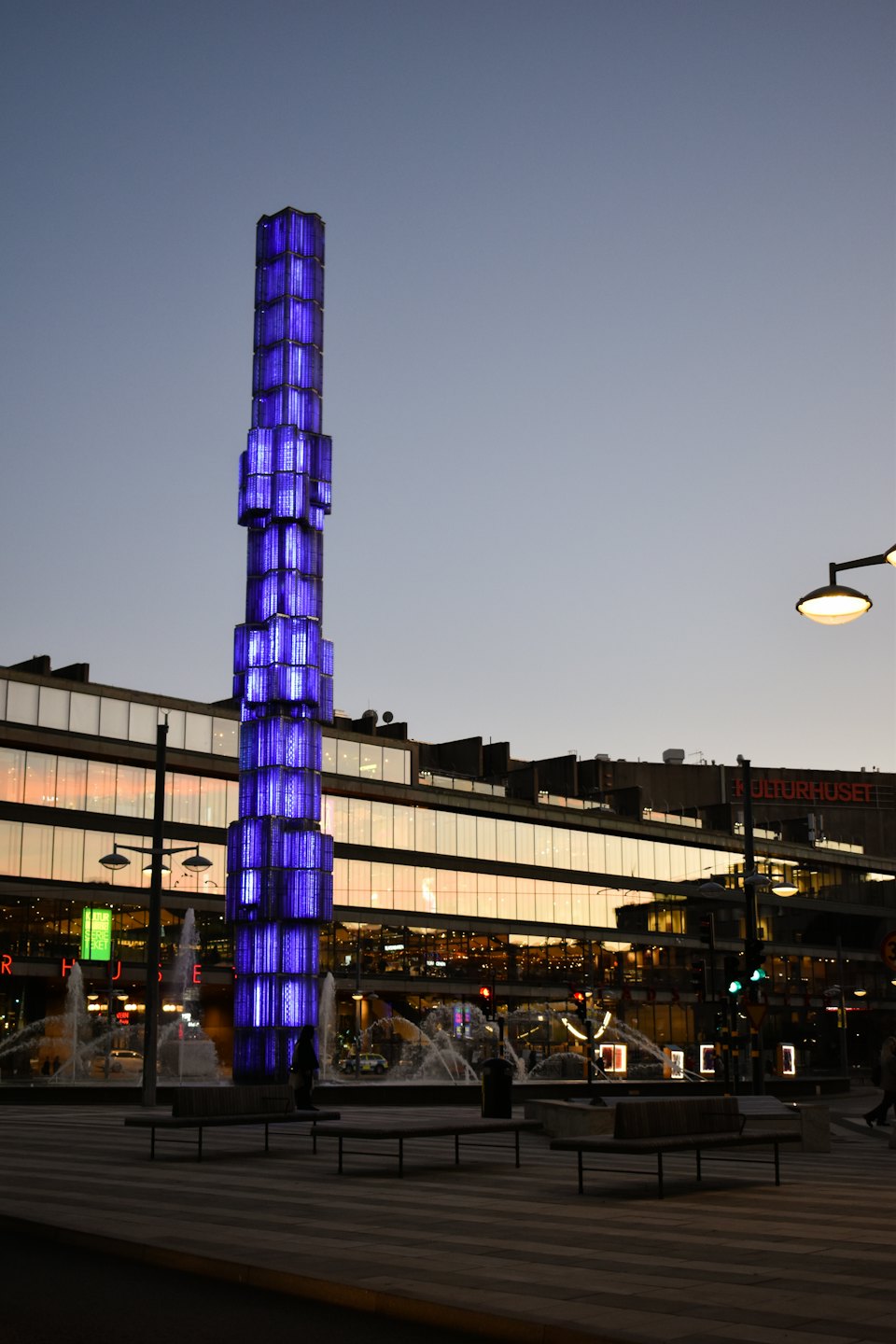 A blue illuminated sculpture stands in a city plaza at dusk.
